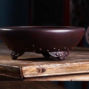 Könnte beinhalten: Ein maroonfarbener Bonsai-Topf aus Keramik mit einer breiten, flachen Schalenform. Der Topf hat drei verzierte Füße und ein dekoratives Band aus kleinen, erhabenen Punkten am Rand. Der Topf steht auf einer Holzoberfläche.