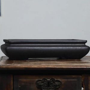 Könnte beinhalten: Ein rechteckiger, schwarzer Bonsai-Topf mit abgerundeten Seiten und einem leicht erhöhten Rand. Der Topf besteht aus einem dunklen Material und steht auf einer Holzoberfläche. Das Design ist schlicht und elegant, geeignet für kleine Pflanzen.