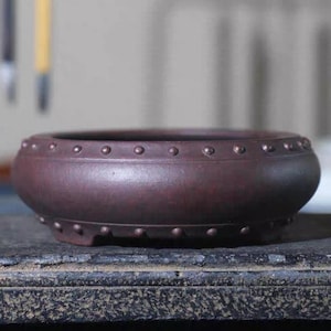 Peut inclure: Un pot en céramique brun foncé de forme arrondie avec un bord décoratif de petits points en relief espacés uniformément.