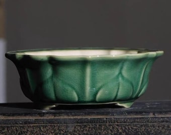 May include: A green ceramic planter with a leaf-like design. The planter has a wide, shallow bowl shape with a white interior and three small feet. The exterior is a glossy, jade green color.