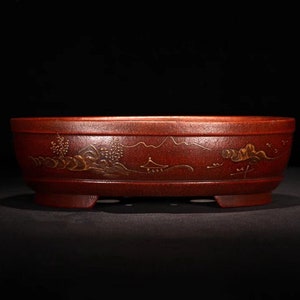 Puede incluir: Una maceta de bonsái ovalada de color rojo con un diseño de paisaje pintado en oro. La maceta tiene tres patas cortas.