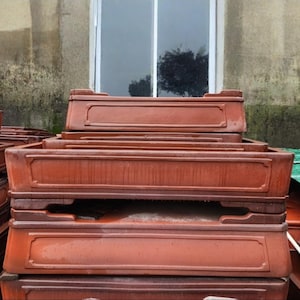 Peut inclure: Plusieurs jardinières rectangulaires brun-rouge sont empilées. Chaque jardinière présente un bord décoratif en relief et une forme rectangulaire. Les jardinières sont empilées les unes sur les autres, avec des espaces visibles. Une fenêtre et un mur patiné en arrière-plan.