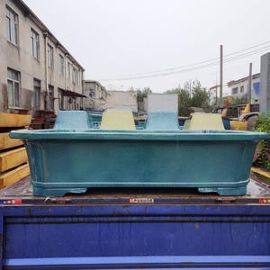 May include: A large, teal-colored rectangular planter with four seat-like structures. The planter is sitting on a blue truck bed, with a building and overcast sky in the background.