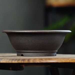 Puede incluir: Una maceta de bonsái de cerámica marrón oscuro con un diseño ancho y poco profundo. La maceta tiene un borde ligeramente ensanchado y una superficie texturizada. Se asienta sobre una base a juego con orificios de drenaje. Ideal para árboles y plantas.