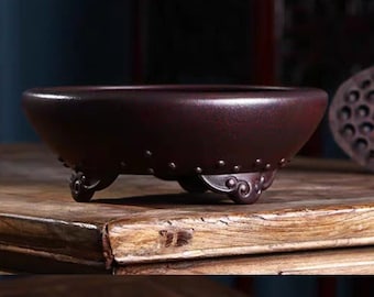 May include: A maroon ceramic bonsai pot with a wide, shallow bowl shape. The pot has three ornate feet and a decorative band of small, raised dots around the rim. The pot is sitting on a wooden surface.