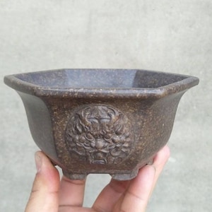 May include: A dark brown octagonal ceramic planter with a raised dragon design on the front. The planter has four small feet and is approximately 12.5 cm in diameter.