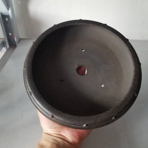 K&ouml;nnte beinhalten: Ein runder, dunkelbrauner Bonsai-Topf aus Keramik mit einem strukturierten Rand. Der Topf hat ein Abzugsloch in der Mitte und mehrere kleinere L&ouml;cher am Rand.