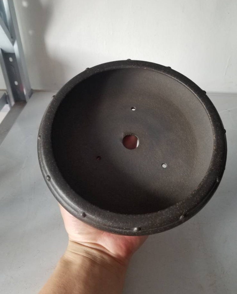 K&ouml;nnte beinhalten: Ein runder, dunkelbrauner Bonsai-Topf aus Keramik mit einem strukturierten Rand. Der Topf hat ein Abzugsloch in der Mitte und mehrere kleinere L&ouml;cher am Rand.