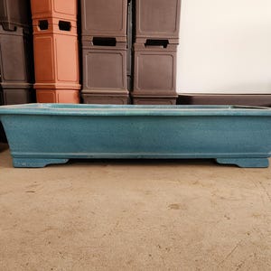 May include: A rectangular, teal-colored bonsai pot with a textured surface. The pot has a slightly flared shape and sits on four small feet. Other pots are visible in the background, suggesting a garden or plant theme.