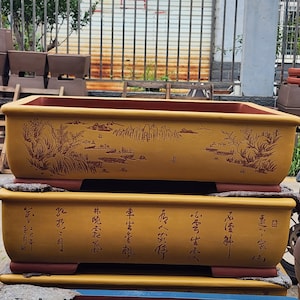 Puede incluir: Dos macetas rectangulares de cerámica marrón amarillento para bonsái. La maceta superior presenta un diseño de paisaje en un color marrón más oscuro. La maceta inferior tiene caracteres chinos. Ambas macetas tienen interior y base rojizo-marrón.