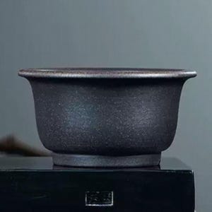 Puede incluir: Una maceta de bonsái de cerámica gris oscuro con forma redondeada y un borde ligeramente acampanado.