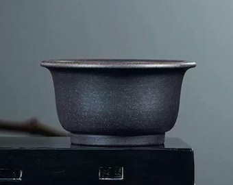 Puede incluir: Una maceta de bonsái de cerámica gris oscuro con forma redondeada y un borde ligeramente acampanado.