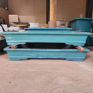 Puede incluir: Dos macetas rectangulares turquesas para bonsái apiladas. Las macetas tienen una superficie texturizada y un diseño ligeramente curvado. Una tabla de madera se coloca dentro de la maceta inferior. Otras macetas turquesas son visibles en el fondo.