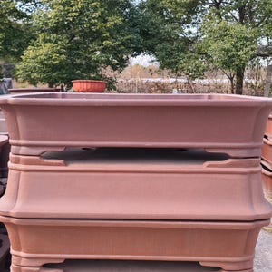 Puede incluir: Una pila de macetas de bonsái de color terracota. Las macetas rectangulares tienen un diseño ancho y poco profundo con un borde ligeramente ensanchado y detalles decorativos. Las macetas están apiladas, mostrando su forma y color.