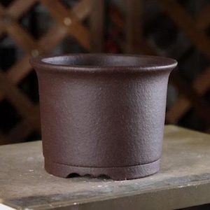 Può includere: Un vaso per bonsai in ceramica marrone scuro con forma arrotondata e un bordo leggermente svasato. Il vaso ha tre piccoli piedini sul fondo.
