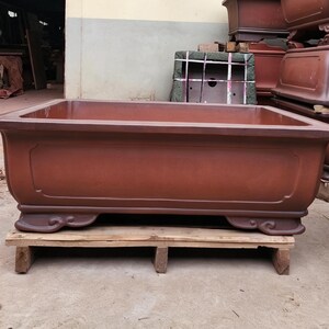 May include: A large, rectangular, reddish-brown ceramic planter with a smooth, matte finish. The planter features a wide, flat rim, a recessed panel design on the front side, and four decorative, curved feet at the base for stability and elevation.