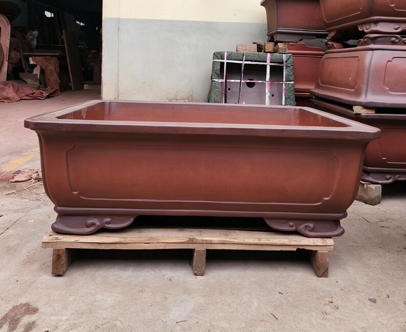 May include: A large, rectangular, reddish-brown ceramic planter with a smooth, matte finish. The planter features a wide, flat rim, a recessed panel design on the front side, and four decorative, curved feet at the base for stability and elevation.