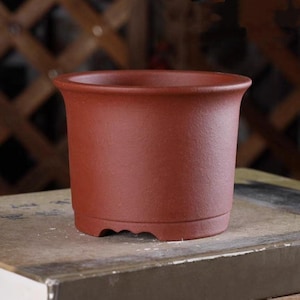 May include: A terracotta colored ceramic pot with a rounded bottom and a slightly flared rim. The pot is sitting on a wooden surface.