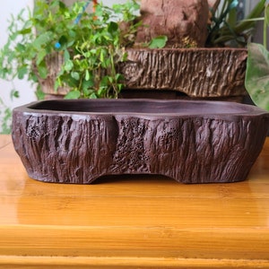 Op de afbeelding: Een rechthoekige bonsai pot van donkerbruin aardewerk met een ruw, getextureerd oppervlak dat lijkt op boomschors. De pot heeft een licht gebogen vorm en een brede basis.