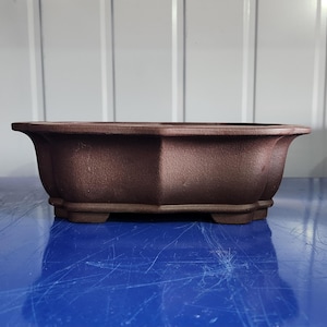 May include: A rectangular, brown bonsai pot with a textured surface. The pot has a wide, shallow design with a slightly flared rim and four small feet. The pot is made of a dark brown material.