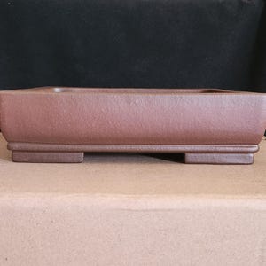 Può includere: Un vaso per bonsai rettangolare in ceramica marrone con una superficie leggermente strutturata. Il vaso ha un profilo basso con angoli arrotondati e poggia su quattro piccoli piedini rettangolari. Lo sfondo è scuro.