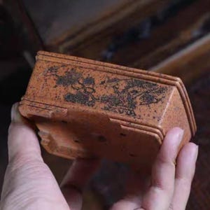 Op de afbeelding: Een kleine, rechthoekige bonsai pot in terracotta kleur met een donker, gestileerd boomontwerp. De pot heeft een getextureerd oppervlak en een trapsgewijs ontwerp. De pot wordt in een hand gehouden.