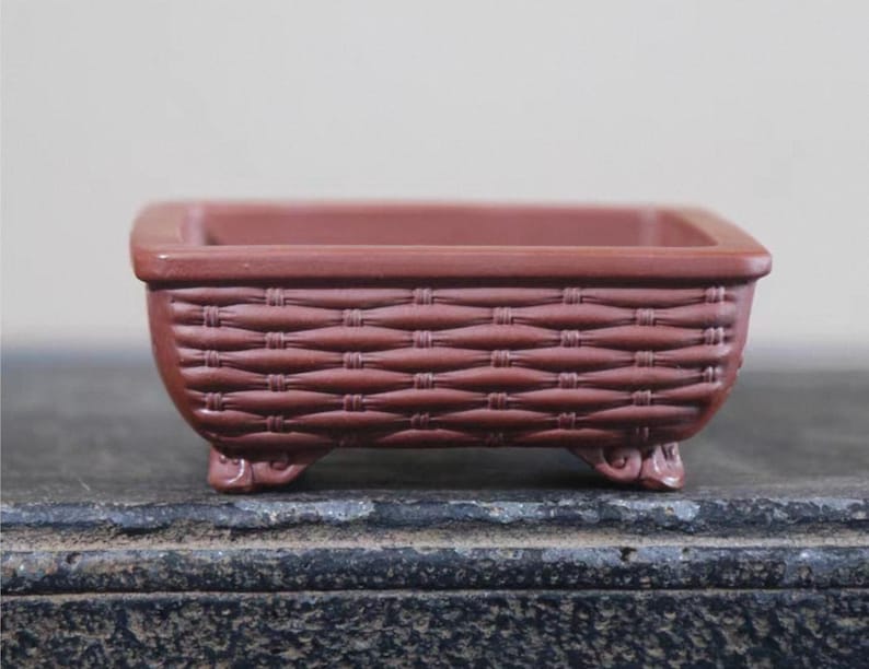 Peut inclure: Petit pot de fleurs rectangulaire en c&eacute;ramique brun rouge&acirc;tre avec un motif de panier tress&eacute;. Le pot a un dessus carr&eacute; et repose sur quatre petits pieds d&eacute;coratifs. Id&eacute;al pour les petites plantes ou les succulentes.