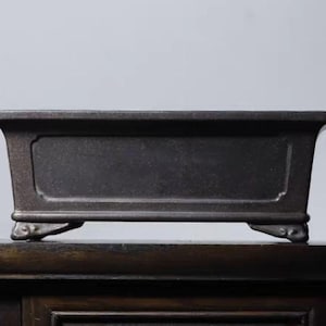 Könnte beinhalten: Ein dunkelbrauner rechteckiger Bonsai-Topf mit leicht gebogenem Rand und dekorativen Füßen. Der Topf hat eine eingelassene Platte auf der Vorderseite und besteht aus einem matten Material. Der Topf ist für den Anbau kleiner Pflanzen konzipiert.