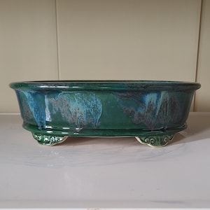 Puede incluir: Maceta de bonsái ovalada de cerámica verde con un esmalte azul y blanco. La maceta tiene tres patas y es perfecta para exhibir plantas pequeñas.