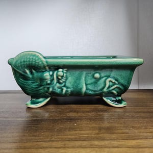 May include: A small, rectangular, ceramic planter in a teal green colour. The planter has a bathtub shape with decorative relief details and sits on three small feet. Suitable for small plants or succulents.