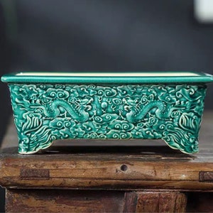 Könnte beinhalten: Ein grüner Keramik-Pflanztopf mit Drachenmotiv. Der Topf hat eine rechteckige Form mit abgerundeten Kanten und vier kleinen Füßen.