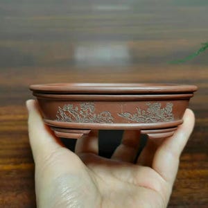 May include: A brown, oval-shaped ceramic bonsai pot featuring a detailed, etched landscape scene on its exterior side. The vessel has a wide, flat rim, a slightly tapered body, and is supported by four small, sturdy feet at the base.