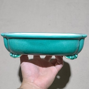 May include: A teal glazed ceramic bonsai pot with an oval shape and four decorative feet.