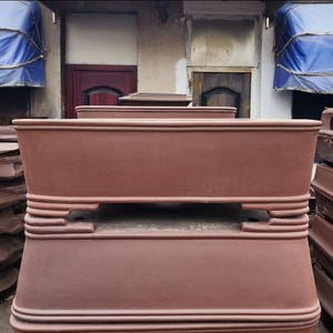 May include: A selection of stacked, rectangular terracotta-coloured planters. The planters have a classic design with a slightly flared base and a decorative band around the middle. The background includes doors and blue awnings.