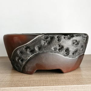 May include: A round ceramic bonsai pot featuring a smooth, dark reddish-brown base and a contrasting textured, charcoal-black band across the upper section. The black portion displays a rough, cratered surface with deep, irregular indentations.