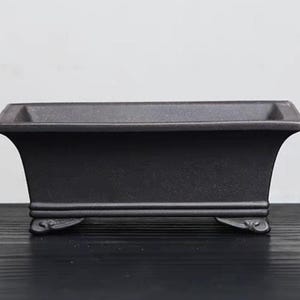 Peut inclure: Un pot rectangulaire en céramique noire avec un bord légèrement évasé et quatre pieds décoratifs. Le pot est fait d'un matériau rugueux et texturé.