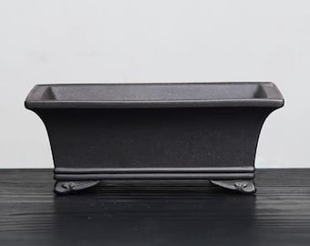 Puede incluir: Un macetero rectangular de cerámica negra con un borde ligeramente acampanado y cuatro patas decorativas. El macetero está hecho de un material rugoso y texturizado.