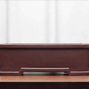 May include: A rectangular, dark brown bonsai pot. The pot has a simple, modern design with a slightly flared shape and a raised base. The pot is made of a matte material and sits on a wooden surface.