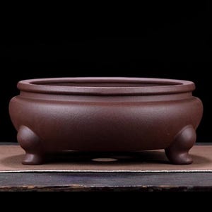 May include: A brown, oval ceramic planter with three rounded feet. The planter has a textured surface and a wide, open top. The dark background accentuates the planter's shape and colour.