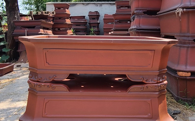 Pode incluir: Uma cole&ccedil;&atilde;o de vasos retangulares para bonsai na cor terracota. Os vasos t&ecirc;m uma base decorativa com volutas e um topo retangular com um painel embutido. Os vasos s&atilde;o de v&aacute;rios tamanhos e formas, adequados para bonsai.