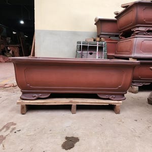 May include: A large, rectangular, reddish-brown ceramic bonsai pot with a smooth finish, featuring a recessed panel design on the front and decorative, curved feet at the base. The pot is positioned on a wooden pallet in an industrial setting.