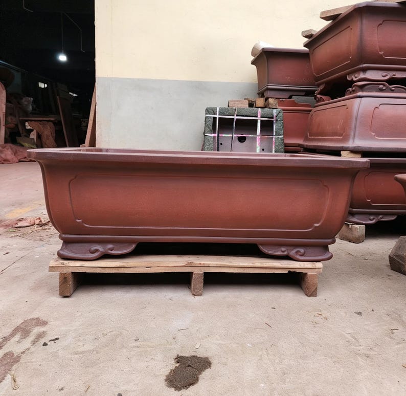 May include: A large, rectangular, reddish-brown ceramic bonsai pot with a smooth finish, featuring a recessed panel design on the front and decorative, curved feet at the base. The pot is positioned on a wooden pallet in an industrial setting.