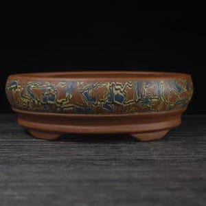 Könnte beinhalten: Ein brauner Bonsai-Topf aus Keramik mit einem dekorativen Muster in Blau und Gelb. Der Topf ist oval geformt, hat ein niedriges Profil und drei kleine Füße. Der Rand weist ein detailliertes Design auf.