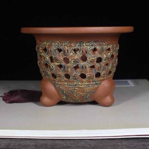 May include: A terracotta-colored ceramic planter with a decorative, openwork design around the middle. The planter has a wide rim and is supported by three rounded feet. The openwork pattern features a mix of brown, blue, and yellow hues.