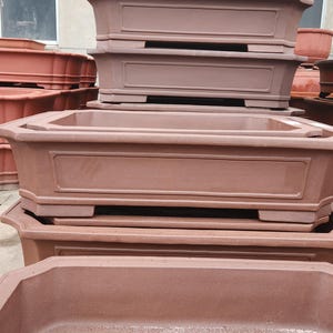 Puede incluir: Una colección de macetas para bonsái rectangulares y octogonales apiladas, de color marrón rojizo. Las macetas tienen una superficie texturizada y paneles decorativos. La imagen muestra varios tamaños y formas, adecuadas para el cultivo de bonsáis. Las macetas son de material duradero.