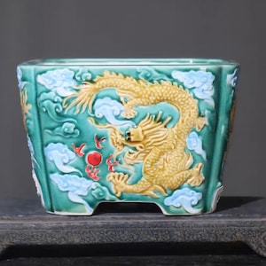 May include: A square, turquoise ceramic planter with a detailed relief of a golden dragon amidst clouds. The dragon is the focal point, with red accents and light blue clouds. The planter sits on a dark, ornate stand.