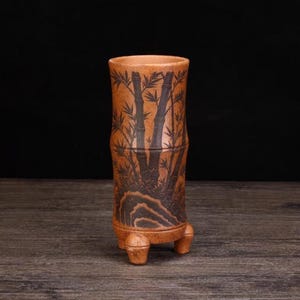 Könnte beinhalten: Eine zylindrische, braune Keramikvase mit Bambusdesign. Die Vase hat eine strukturierte Oberfläche und wird von drei kleinen Füßen getragen. Das Bambusdesign ist schwarz und bedeckt den Großteil der Vasenoberfläche.