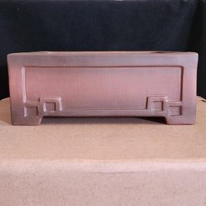 Könnte beinhalten: Ein rechteckiger, rotbrauner Bonsai-Topf aus Keramik. Der Topf hat einen erhabenen rechteckigen Rand und dekorative quadratische Akzente an den Ecken. Der Topf steht auf einer Kartonoberfläche.