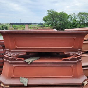 Puede incluir: Una pila de macetas de bonsái rectangulares de cerámica marrón rojizo. Cada maceta tiene un borde decorativo en relieve y detalles ornamentados. Las macetas están apiladas al aire libre bajo un cielo nublado.