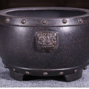 Könnte beinhalten: Ein dunkelgrauer, runder Keramik-Pflanztopf mit strukturierter Oberfläche. Der Pflanztopf hat ein dekoratives Gesichts-Emblem und ist mit Metallnieten am Rand und am Boden verziert. Der Pflanztopf steht auf einem quadratischen Sockel.